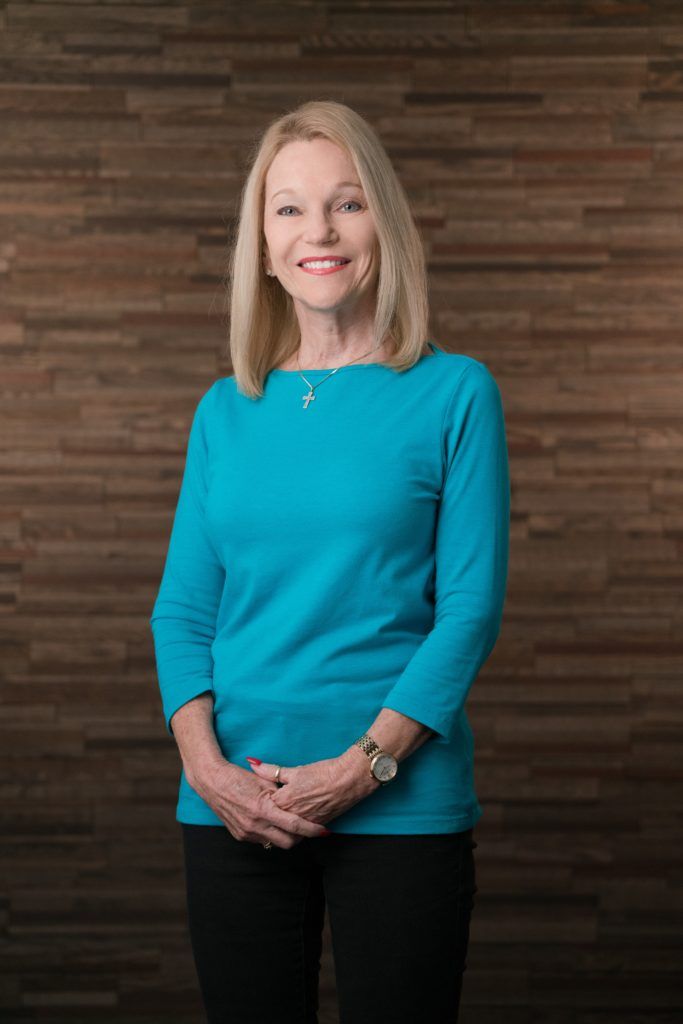 a woman stands in a blue shirt in front of a wooden background for her company headshot. 