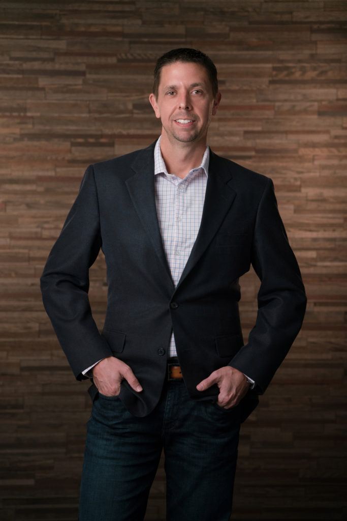 a man stands with his hands in pockets in front of a wooden background for her company headshot. 