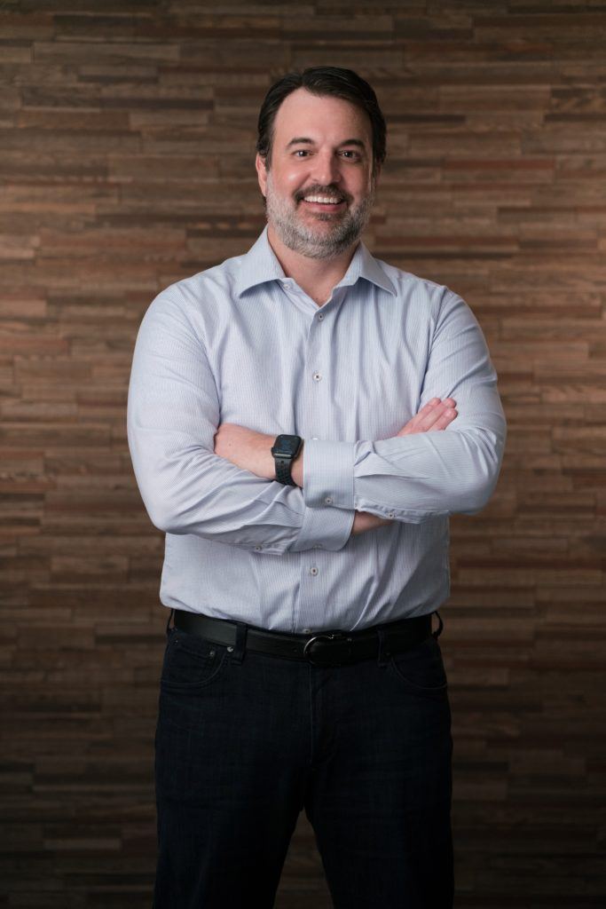 A man stands in a light colored button down shirt with his arms crossed over his chest in front of a wooden background for his company headshot. 