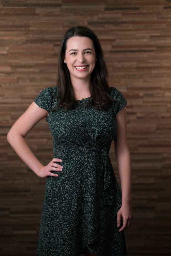 A woman smiles, standing in a black and white polka dot dress, with one hand on her hip in front of a wooden backdrop for her company headshot. 